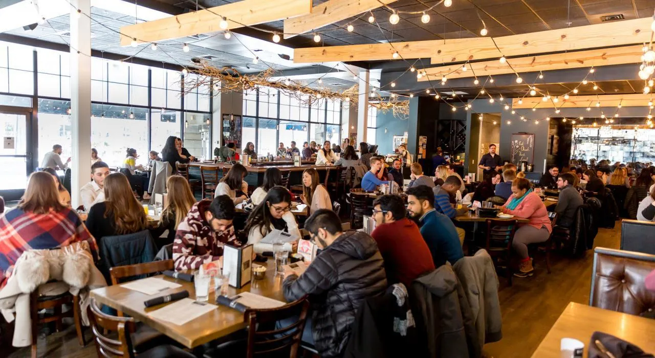 A crowded, brightly lit modern restaurant dining room filled with people eating at tables A crowded, brightly lit modern restaurant dining room filled with people eating at tables